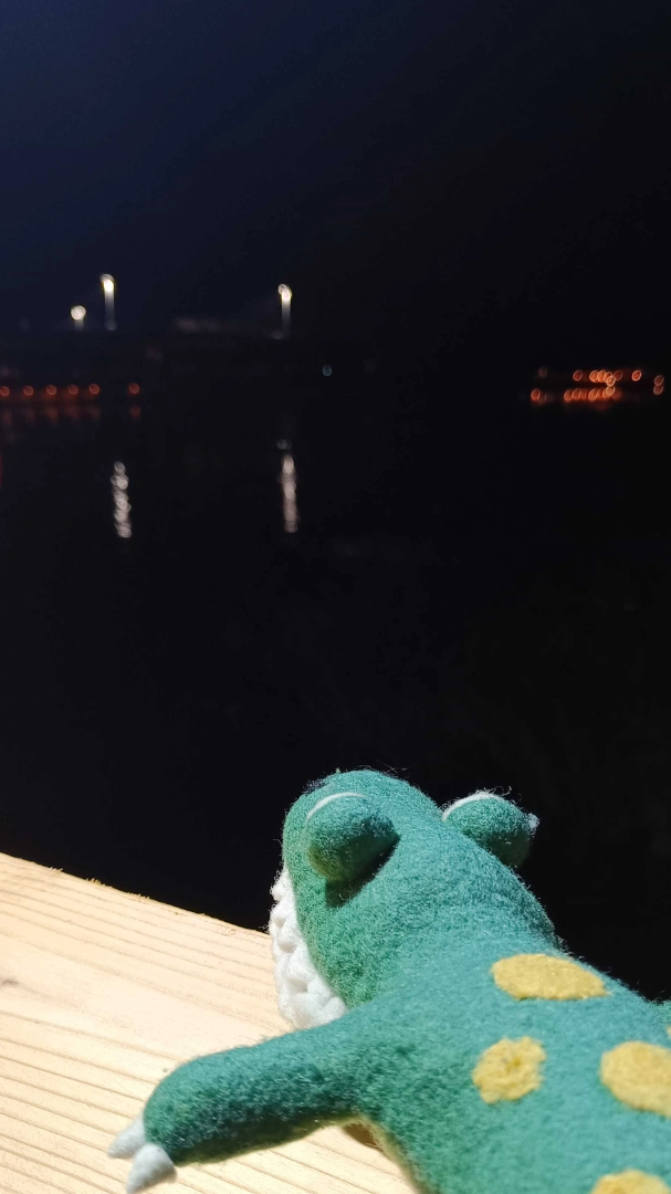 A crocodile plush with yellow spots on his back looking out towards the river at night.